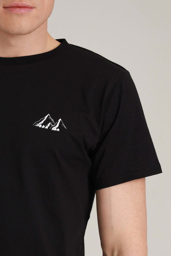 Close Up Foto von schwarzem Shirt  mit Berge-Motiv (weißer Druck). Das Berge-Motiv befindet sich über der linken Brust des Shirt-Trägers. Das Motiv besteht aus 3 Bergen: 
von links nach rechts: beginnend mit einem großen Berg, daneben versetzt nach vorne ein kleiner Berg und ein großer Berg rechts daneben. Die Berge sind alle verbunden. Das kleine Extra sind die winzigen Bäume (Tannen), die sich am Fuß der Berge befinden. Die Berge sind schattiert, sodass Tiefe und Kontur entsteht. 