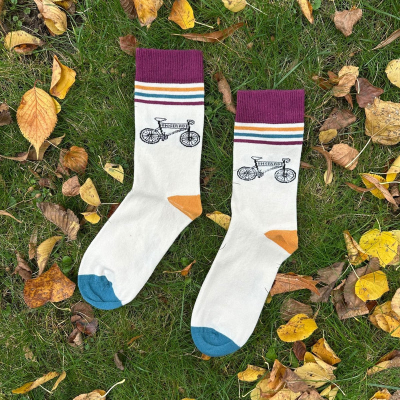 Bike Socke – Nachhaltige Bio-Socken mit Bike-Motiv. Die Socken liegen im grünen Gras, umgeben von Herbstblättern. Der Sockenbund ist ca. 4 cm breit, dunkelrot. Darunter befinden sich 3 Querstreifen (ca 3-5mm dick) in Ockergelb, dunkelgrün, dunkelrot. Darunter liegt das gezeichnete Bike-Motiv. in der Querstange des Bikes steht "Ungerade". Die Ferse ist Ockergelb und die Sockenspitze dunkelrgün.- - Ungerade Wien
